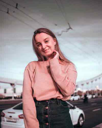 A photo of a beautiful Russian woman in a pink sweater standing on the side of the road, with a white car passing behind her